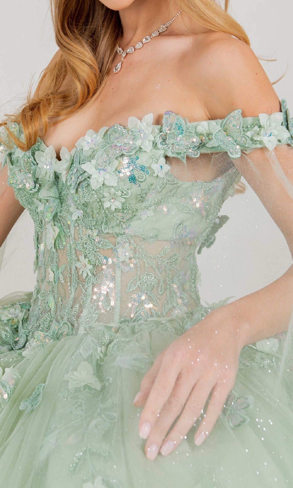 A woman in the Cinderella Couture 8075J quinceanera dress—light green, off-the-shoulder, with 3D floral and butterfly appliqués, sequins, tulle, a detachable sheer cape—and a necklace, her wavy blonde hair styled down.